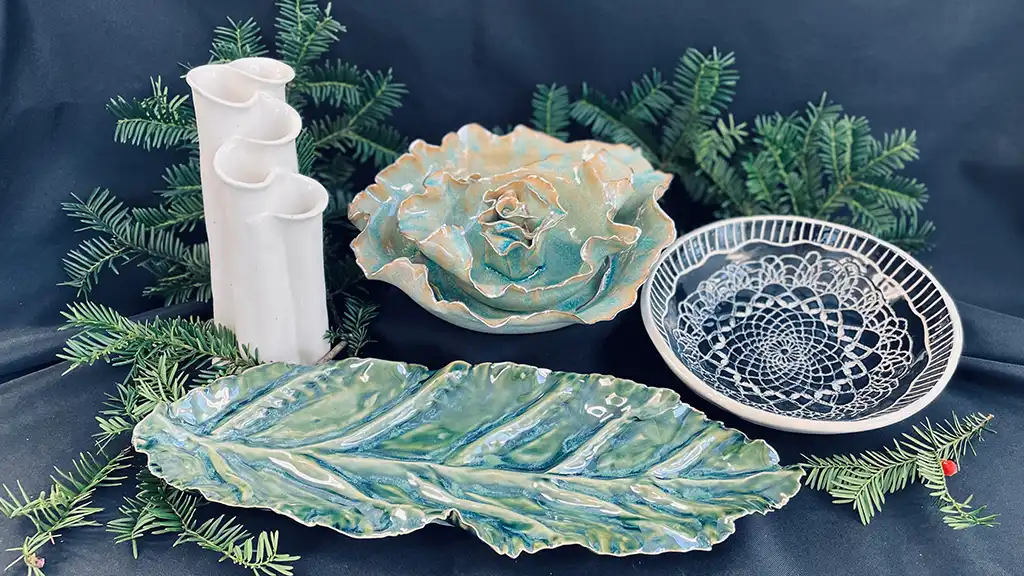Handcrafted ceramics including leaf platters and ruffled bowls displayed with pine boughs